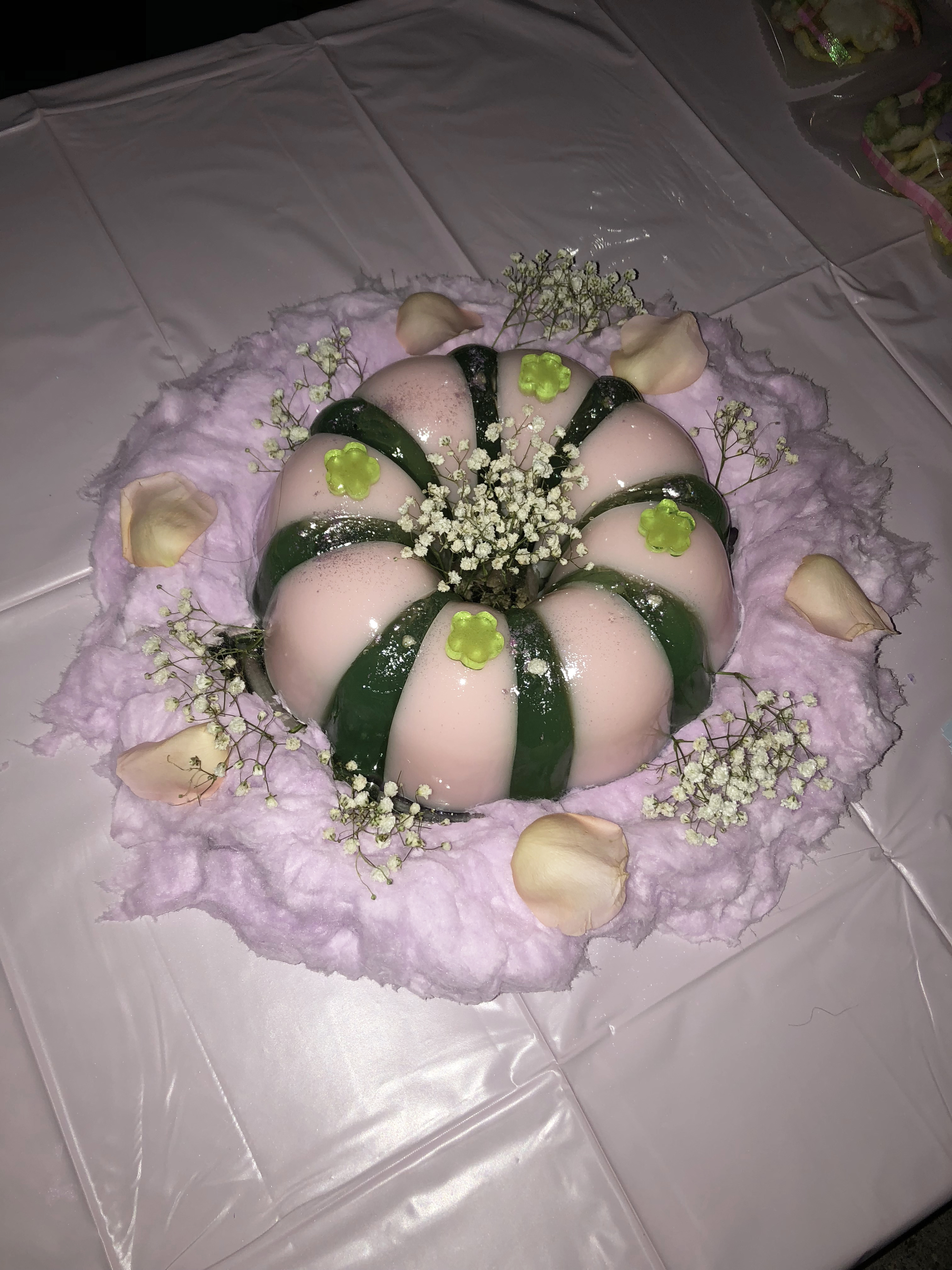 A pink and green jello cake surrounded by cotton candy on a pink table